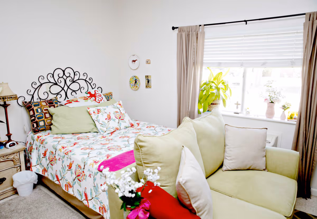 A cozy bedroom featuring a bed with a decorative wrought iron headboard, floral patterned bedding, and multiple pillows. Next to the bed is a wooden nightstand with a lamp and a small trash bin on the floor. In front of the bed is a light green loveseat with cushions. A window with beige curtains and white blinds lets in natural light, and there are several potted plants and small decorative items on the windowsill. The walls are white with a few small framed pictures hanging.