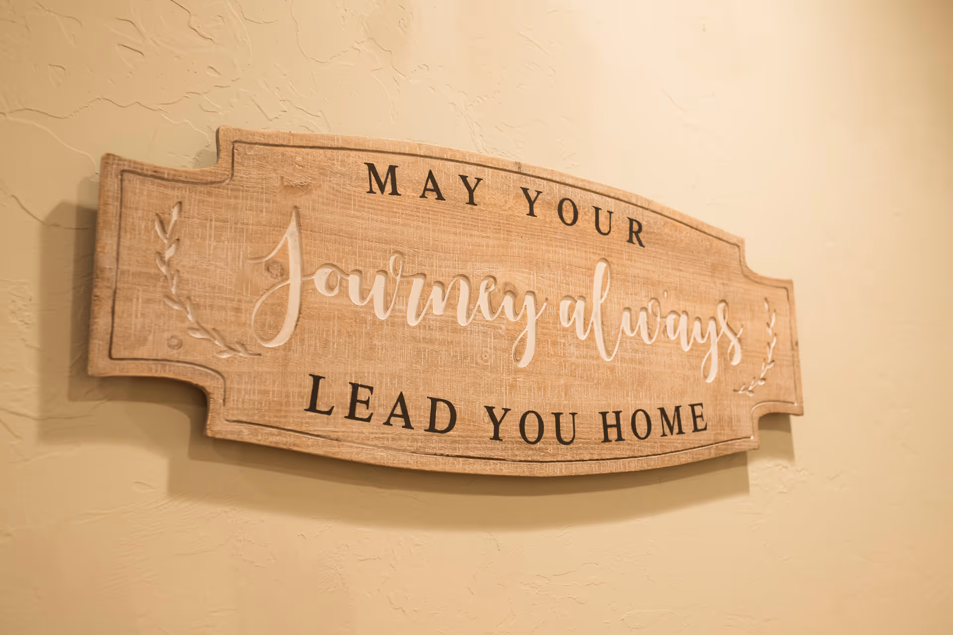 A wooden wall plaque with the inscription 'May your journey always lead you home' mounted on a textured beige wall.