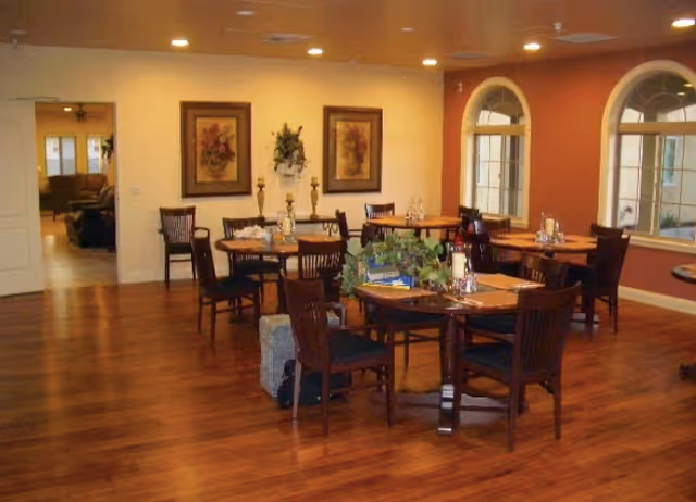 A dining room with several round wooden tables and chairs arranged on a polished wooden floor. The tables are set with placemats, glassware, and decorative centerpieces. The room has warm lighting from recessed ceiling lights, two framed floral paintings on the wall, and three large arched windows letting in natural light. An open doorway leads to another room with seating visible.