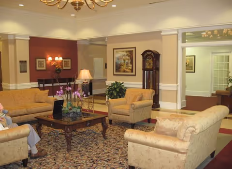 A cozy living room area with beige upholstered sofas and armchairs arranged around a wooden coffee table with a floral centerpiece. The room features a patterned carpet, a grandfather clock, framed artwork on the walls, and warm lighting from table and wall lamps. The walls are painted in neutral tones with white trim, and there is an open doorway leading to another room.