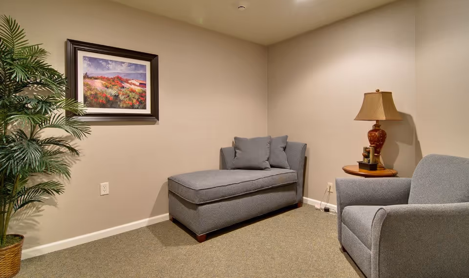 A cozy seating area in a senior living facility with a gray chaise lounge and matching armchair, a wooden side table with a decorative lamp, a framed landscape painting on the wall, and a potted plant in the corner.