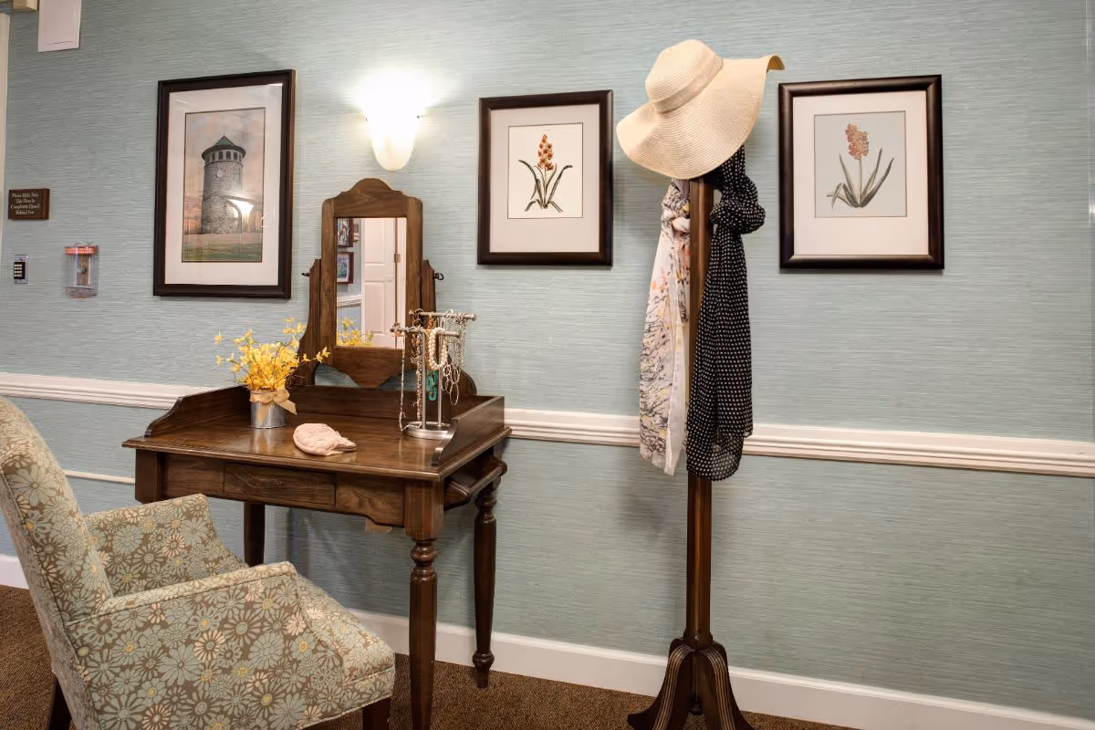 A small wooden vanity with a mirror and floral-upholstered chair sits against a framed-art-covered wall next to a coat rack holding hats and scarves.