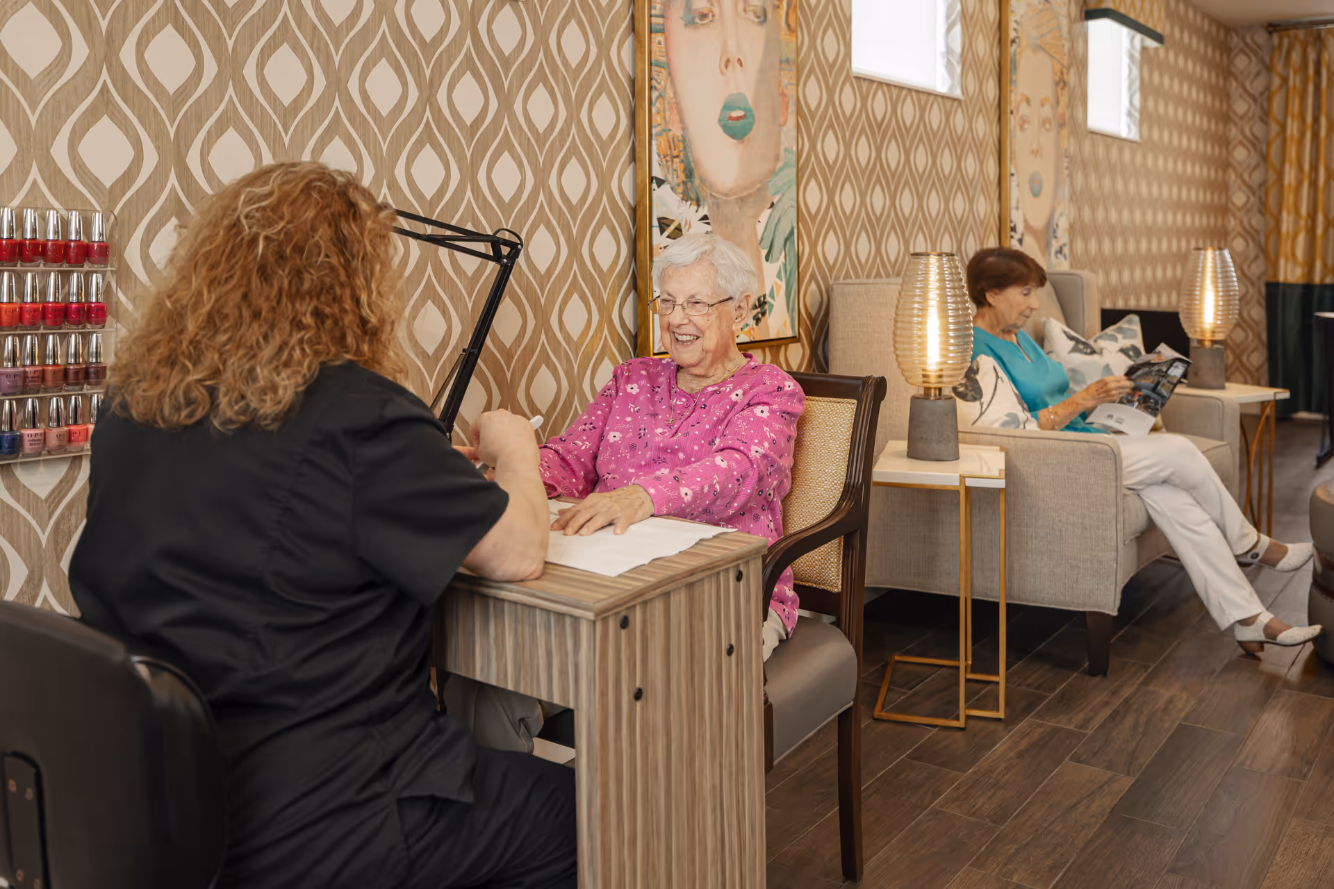 An elderly woman in a pink sweater is smiling while getting a manicure from a technician in a black uniform at a nail station. In the background, another elderly woman in a turquoise top and white pants is seated on a beige armchair reading a magazine. The room has patterned wallpaper, modern lamps on side tables, and framed artwork on the walls.