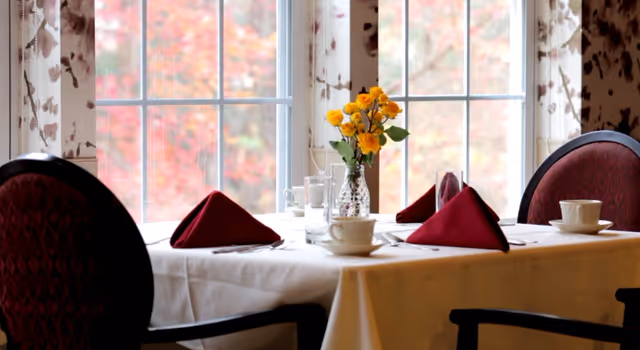 A dining table set for four with white tablecloth, red folded napkins, white cups and saucers, and a vase of yellow flowers in front of a window with sheer floral curtains and a view of autumn foliage outside.