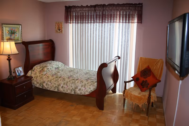 A cozy bedroom with a wooden sleigh bed covered in a floral bedspread, a wooden nightstand with a lamp and framed photos, a chair draped with a yellow throw and a red decorative pillow, vertical blinds covering a large window, and a flat-screen TV mounted on the wall.