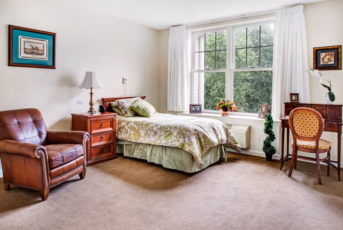 A cozy bedroom in a senior living facility with a single bed covered in floral bedding, a wooden nightstand with a lamp, a brown leather armchair, a wooden desk with a chair, and a large window with white curtains showing green trees outside.