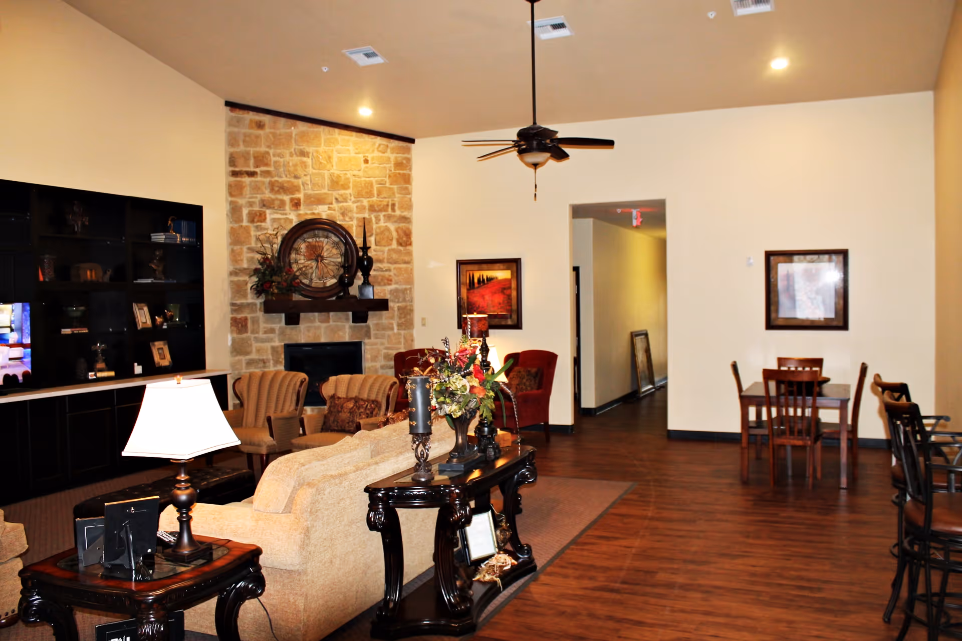 A cozy living room area in an assisted living facility featuring a beige sofa, two armchairs, a stone fireplace with decorative items on the mantel, a ceiling fan, wooden flooring, and a small dining table with chairs in the background.