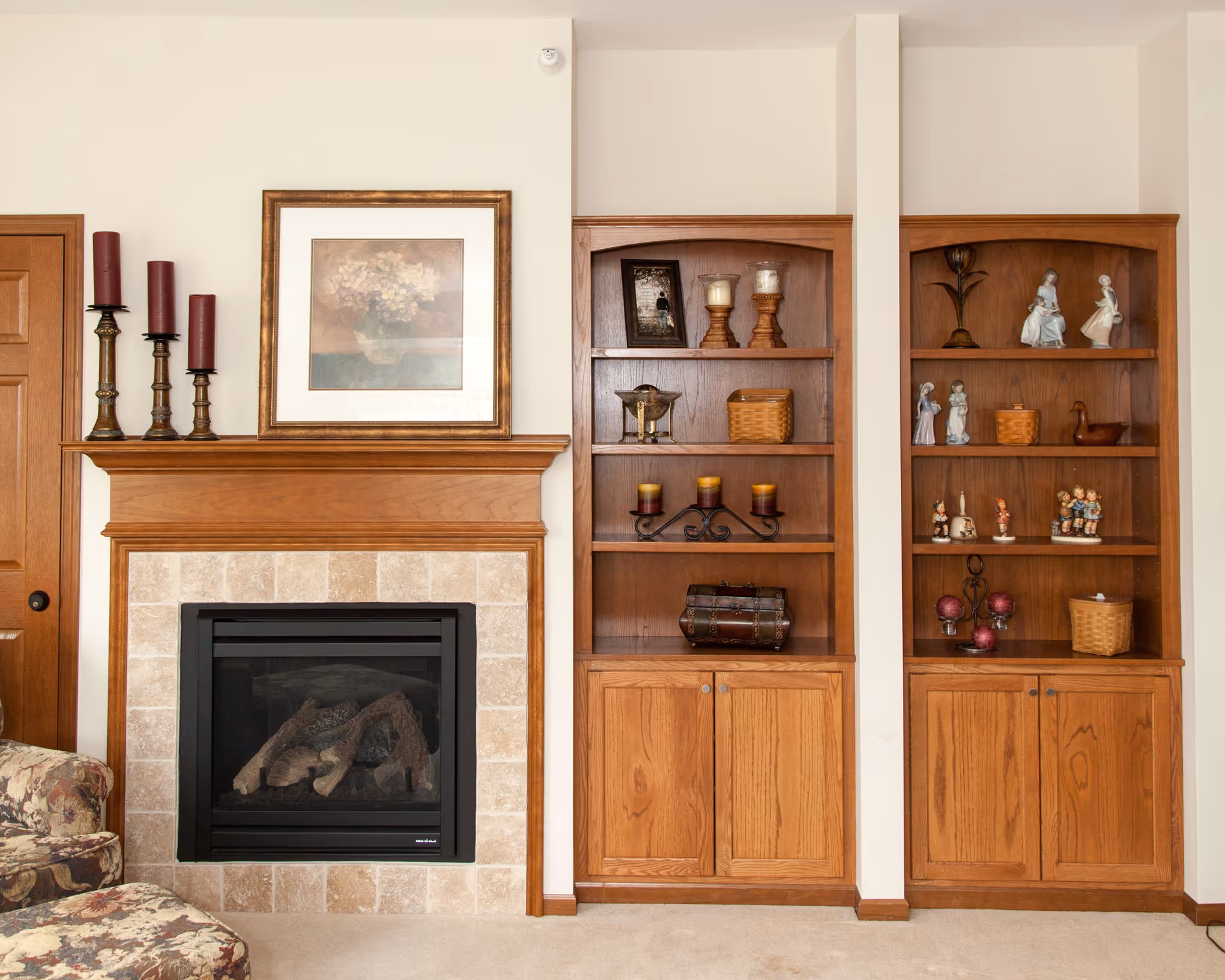 A cozy living room area featuring a wooden fireplace with a tiled surround. Above the fireplace is a framed floral painting and three tall red candles on brass candle holders. To the right of the fireplace are two wooden built-in bookshelves displaying various decorative items including candles, figurines, baskets, and small sculptures. A floral upholstered armchair is partially visible on the left side.