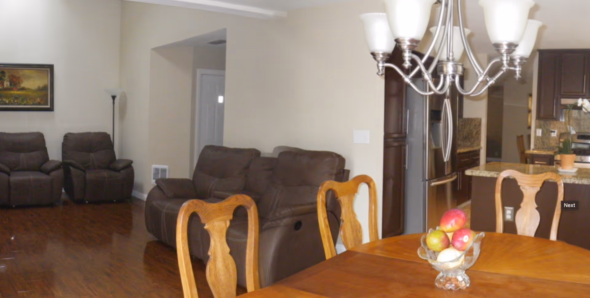 Interior view of a senior living facility showing a dining area with a wooden table and chairs, a bowl of fruit on the table, a chandelier overhead, and a living room area with brown leather recliners and a sofa. The kitchen with dark cabinets and granite countertops is visible in the background.