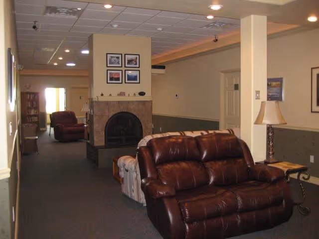 Interior view of a senior living facility common area with brown leather and fabric sofas, a fireplace with framed pictures above it, a side table with a lamp, and a hallway leading to other rooms.