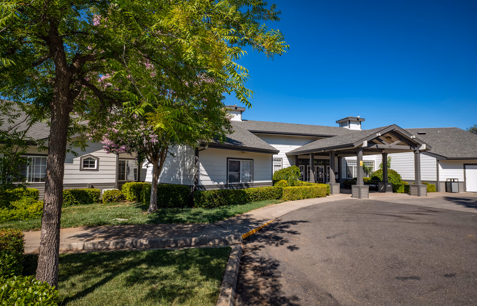 Exterior view of Country Crest Post-Acute facility showing a single-story building with white siding and gray roof under a clear blue sky. There is a covered entrance with columns, surrounded by green bushes and trees with some pink flowers. A paved driveway curves in front of the entrance.