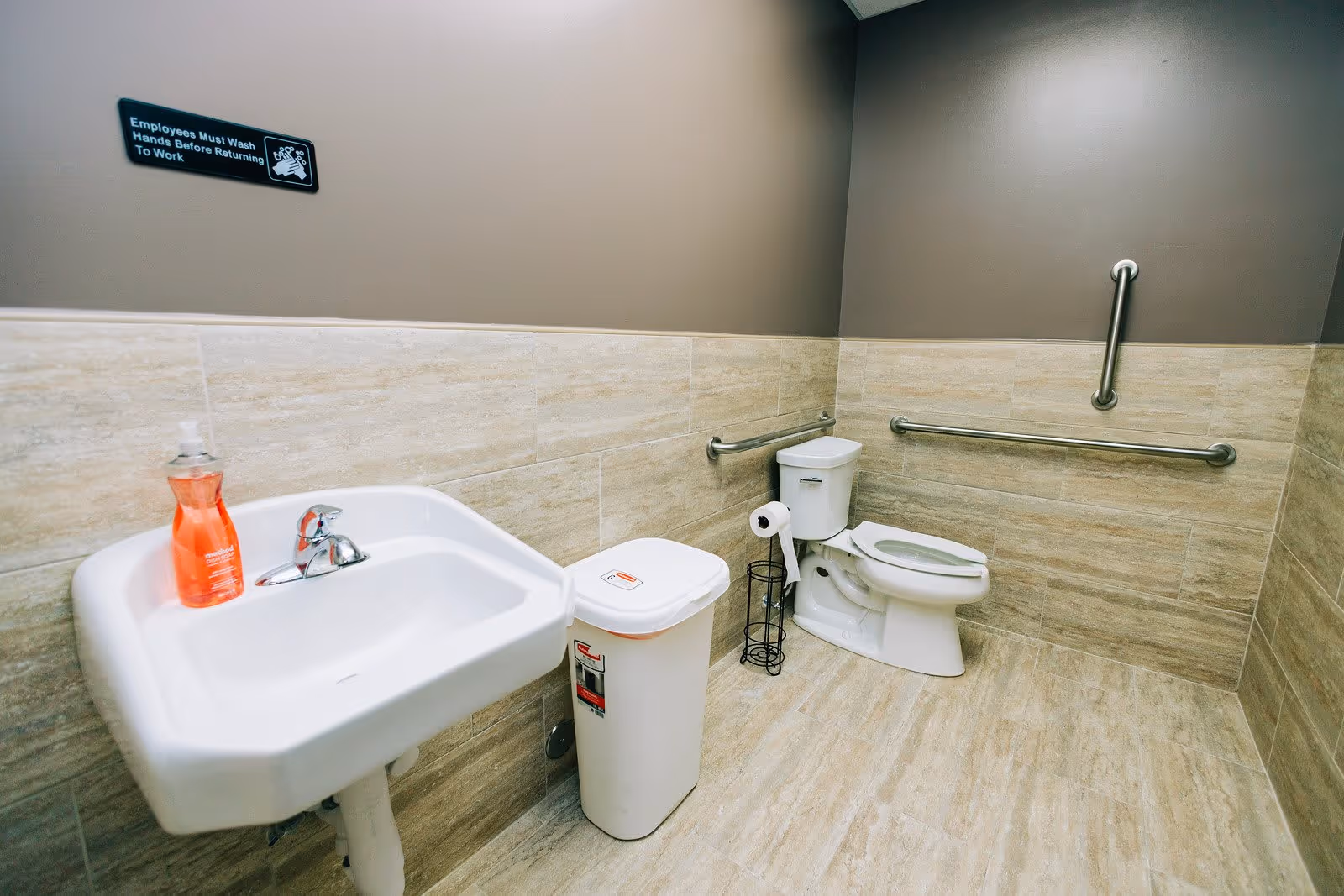A clean and accessible bathroom with beige tiled walls and floor. The room features a white sink with a bottle of orange soap, a white trash bin, a toilet with a toilet paper holder, and multiple stainless steel grab bars on the walls for support. A sign above the sink reads 'Employees Must Wash Hands Before Returning To Work.'