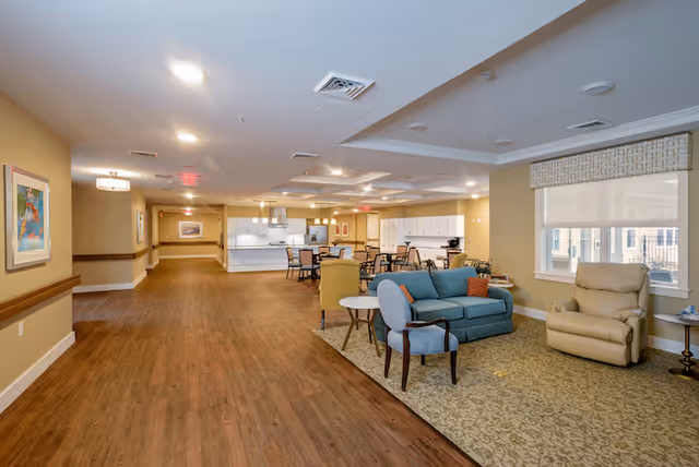 A spacious and well-lit common area in a senior living facility featuring a seating area with a blue sofa, beige recliner, and chairs on a patterned carpet. The background shows a dining area with tables and chairs, and a kitchen with white cabinets and countertops. The walls are painted beige with framed artwork, and large windows allow natural light to enter.