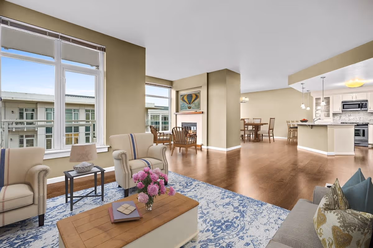 A spacious and bright living area in a senior living facility named Mirabella, featuring large windows with views of an adjacent building, beige walls, and wooden flooring. The room includes two beige armchairs with striped accents, a coffee table with a vase of pink flowers and books, a side table with a lamp, a blue and white patterned rug, a gray sofa with decorative pillows, a fireplace with a hot air balloon painting above it, a dining area with a wooden table and chairs, and a modern kitchen with white cabinetry, pendant lights, and stainless steel appliances.