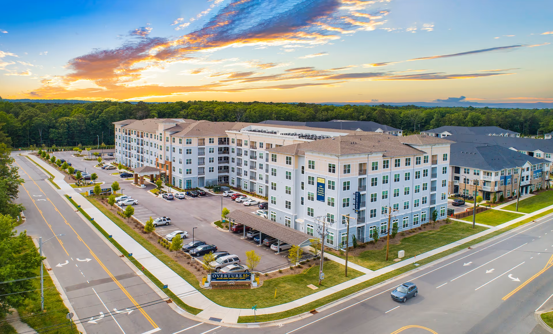 Aerial view of a multi-story Overture Chapel Hill senior living building with parking lot, landscaped grounds, and surrounding roads at sunset.