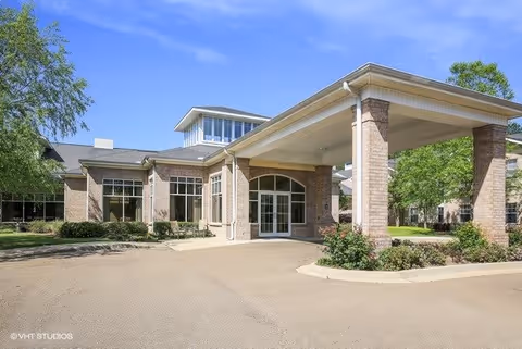 Exterior view of a single-story brick building with large windows and a covered entrance supported by brick columns. The building is surrounded by greenery and trees under a clear blue sky.