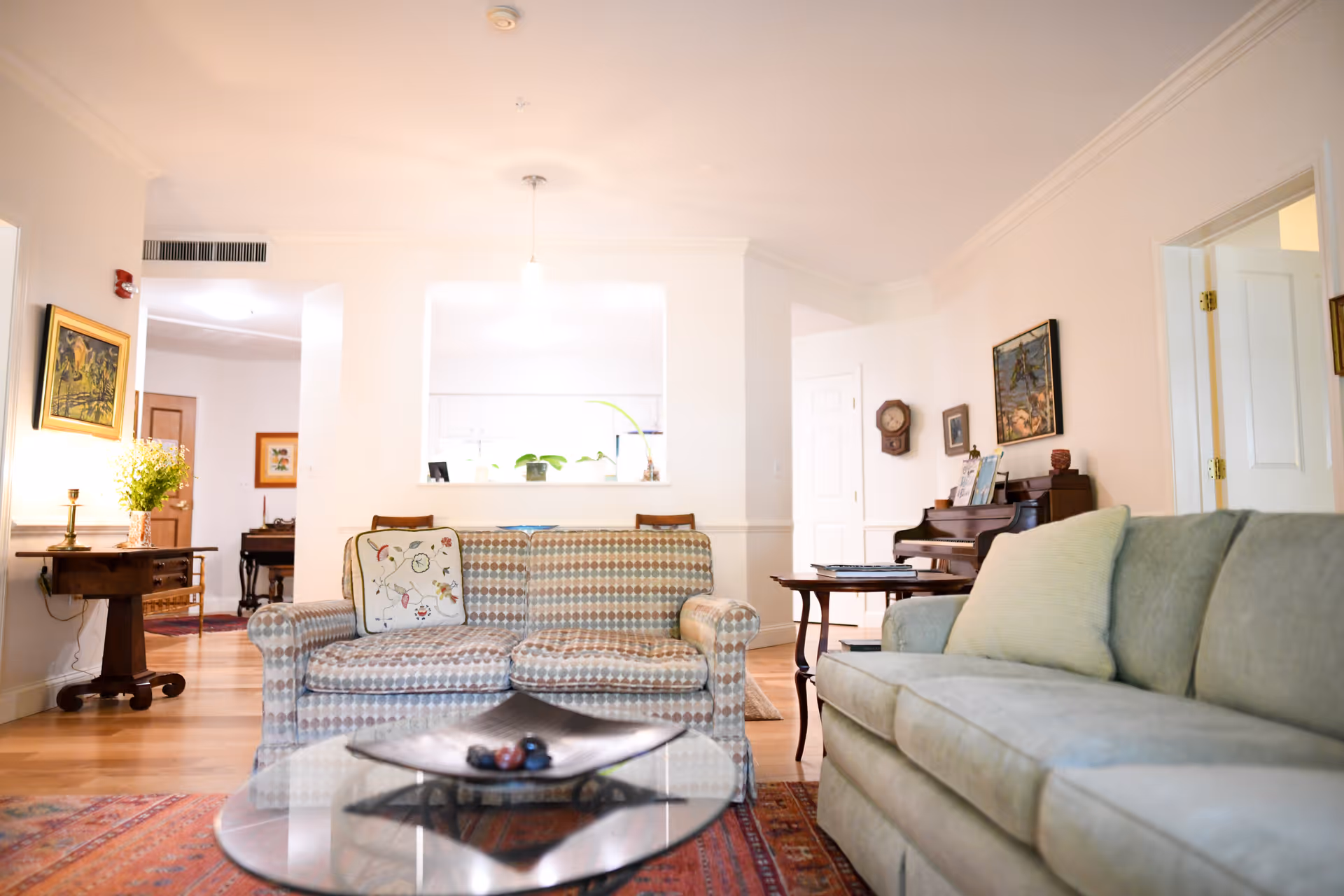 A bright and cozy living room with two sofas facing each other, a glass coffee table with decorative items, a wooden side table with a vase of flowers, and a piano against the wall. The room has light-colored walls, wooden flooring, and several framed paintings and a clock on the walls.