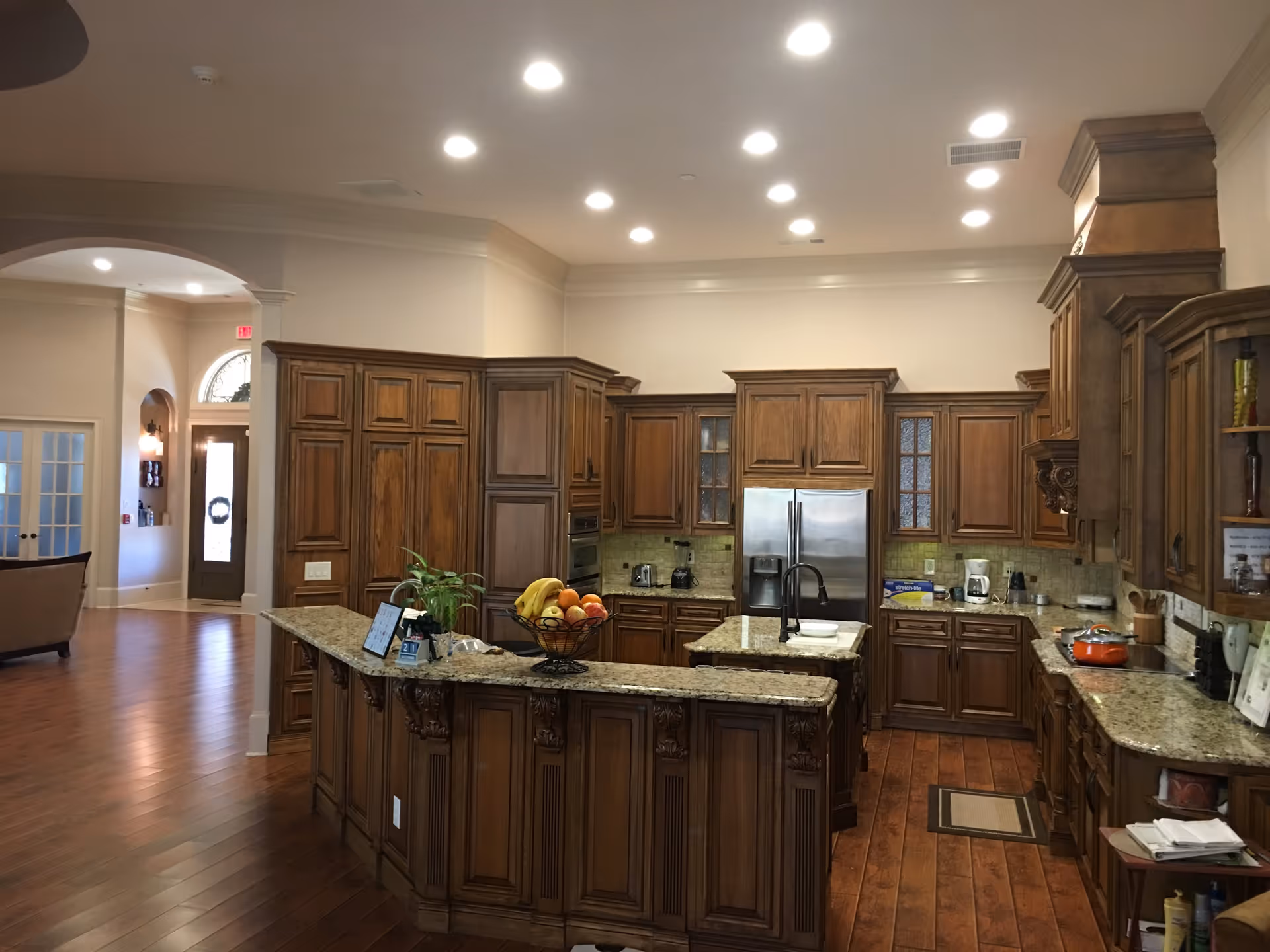 Spacious kitchen area with wooden cabinetry and granite countertops. The kitchen features a large island with a sink, a stainless steel refrigerator, and various kitchen appliances. The floor is wooden, and the ceiling has multiple recessed lights. In the background, there is an open doorway leading to another room with seating and a front door with glass panels.