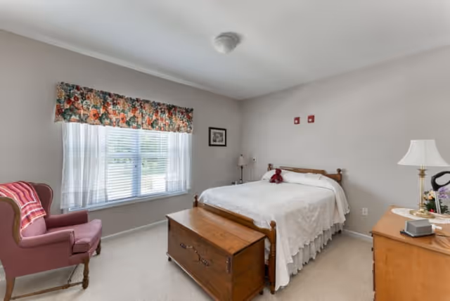 A cozy bedroom with a wooden bed covered in a white bedspread, a wooden chest at the foot of the bed, a wooden dresser with a lamp and flowers, a pink upholstered armchair with a striped blanket, and a window with floral valance and white blinds letting in natural light.