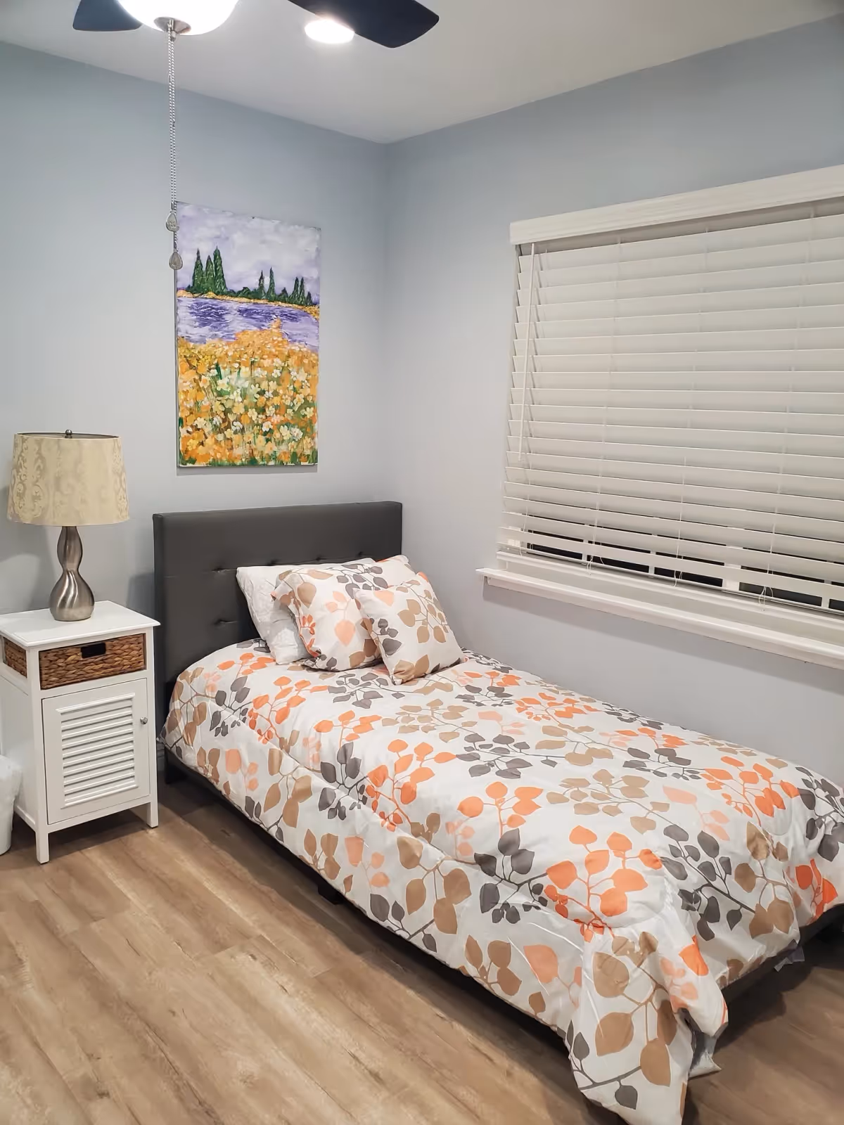 A small bedroom with a single bed featuring a floral patterned comforter and pillows in shades of orange, beige, and gray. Next to the bed is a white nightstand with a wicker drawer and a decorative lamp. Above the bed hangs a colorful painting of a landscape with trees and flowers. The room has light gray walls, wood flooring, a ceiling fan with a light, and a window with white blinds.