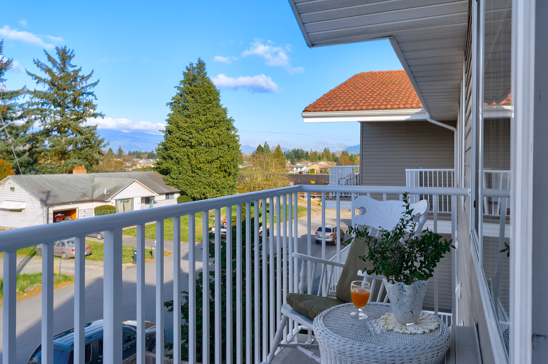 View from a balcony with white railing overlooking a residential neighborhood with houses, trees, and parked cars under a blue sky with some clouds. On the balcony, there is a white wicker table with a potted plant and a glass of orange drink, and a white chair with a cushion.