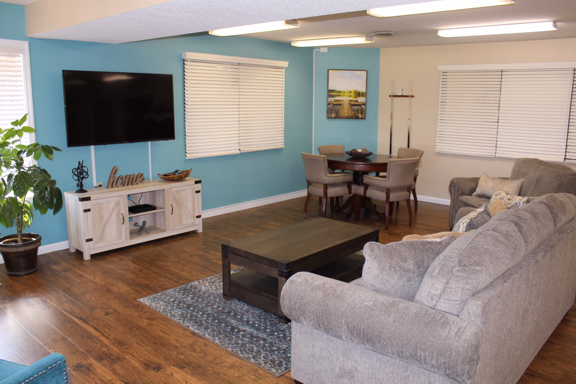 Bright living room with a gray sectional, wooden coffee table, wall-mounted TV, dining table in the background, and a teal accent wall.