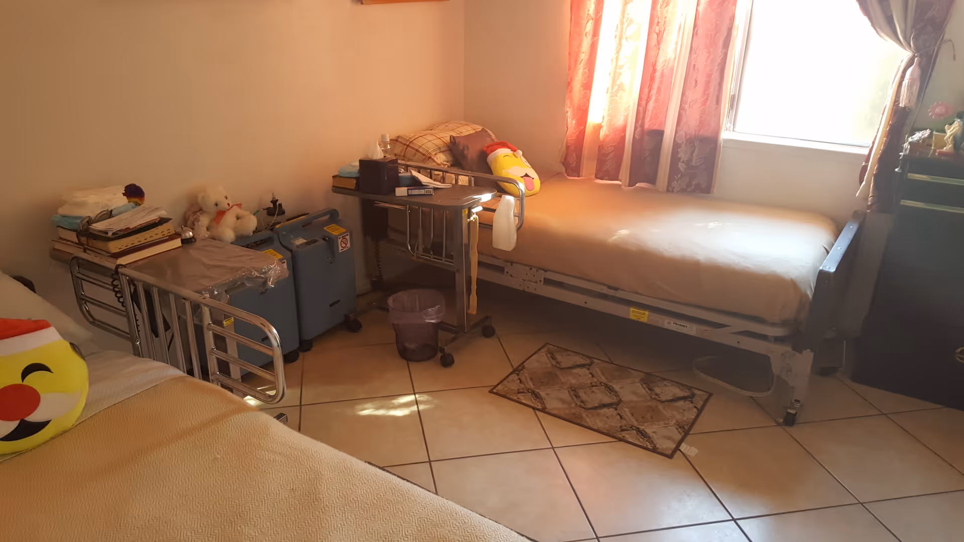 A small room with two hospital-style beds, each with beige bedding and an emoji pillow. There are medical devices and books on a small table between the beds. A window with pink curtains lets in natural light, and a small rug is on the tiled floor.
