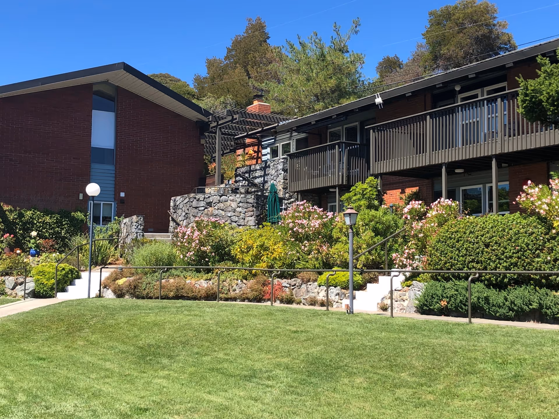Exterior view of a senior living facility with a well-maintained lawn, landscaped garden with various shrubs and flowering plants, and a two-story brick building with balconies and a stone staircase under a clear blue sky.