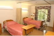 A room with two hospital-style beds covered with pink bedspreads, a wooden bedside table between them, a window with floral curtains, and medical equipment near one of the beds.