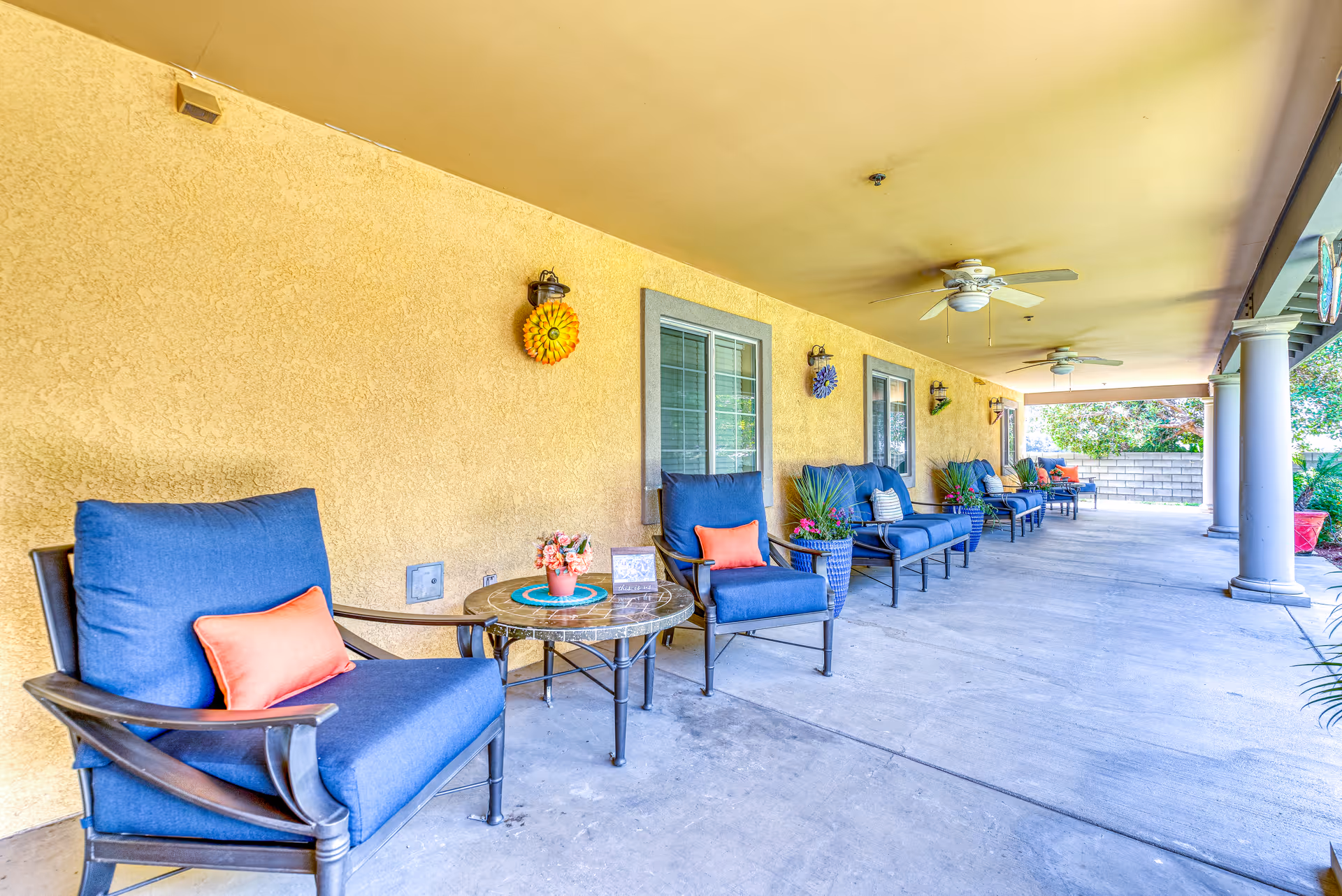 Covered outdoor patio area with several blue cushioned chairs and small tables arranged along a yellow stucco wall. The patio has ceiling fans, decorative wall hangings, potted plants, and columns supporting the roof. The area is spacious and well-lit with natural light.
