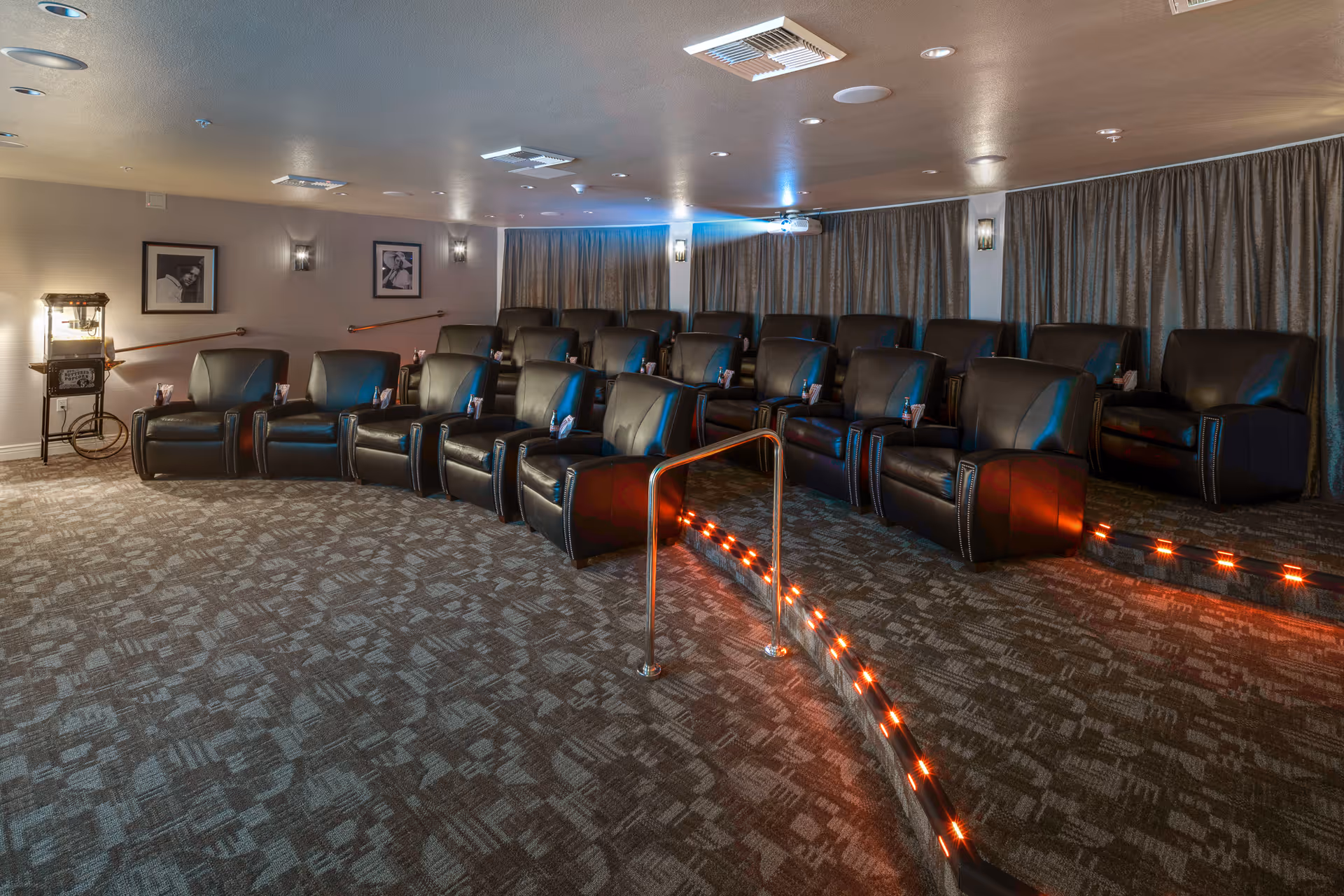 A small theater room with two rows of black leather recliner chairs arranged on a carpeted floor with a handrail and red LED lights along the steps. The walls are light-colored with framed black and white photos and wall sconces. A popcorn machine is visible on the left side of the room.