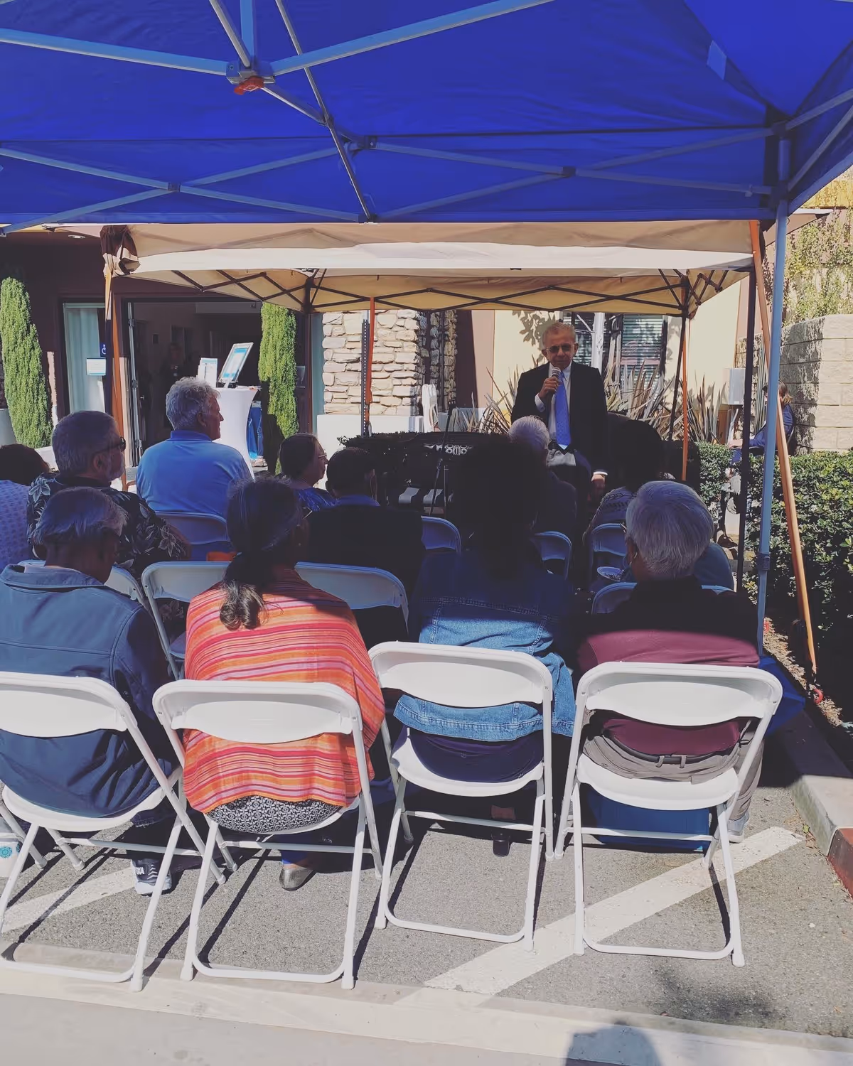 A group of elderly people seated on white folding chairs under a blue canopy, listening to a man in a suit speaking into a microphone outside a building with stone and stucco walls.