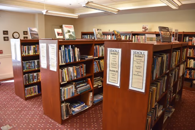 Interior view of a senior living facility library with wooden bookshelves filled with books. The shelves are labeled with sections such as American History, Cooking, and Local Works. The room has a patterned carpet and fluorescent ceiling lights.