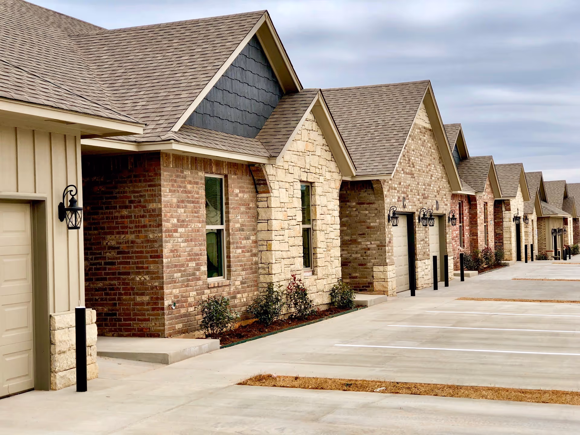 Row of single-story residential units with brick and stone facades, each featuring a garage with black lantern-style lights mounted beside the doors. The units have pitched shingle roofs and small landscaped areas with shrubs in front. The scene is under an overcast sky with a wide concrete driveway and parking spaces in front.
