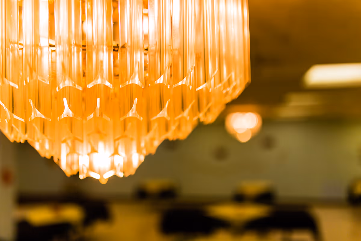 Close-up view of a warm-toned chandelier with blurred tables and chairs in the background, suggesting an indoor dining or gathering area.