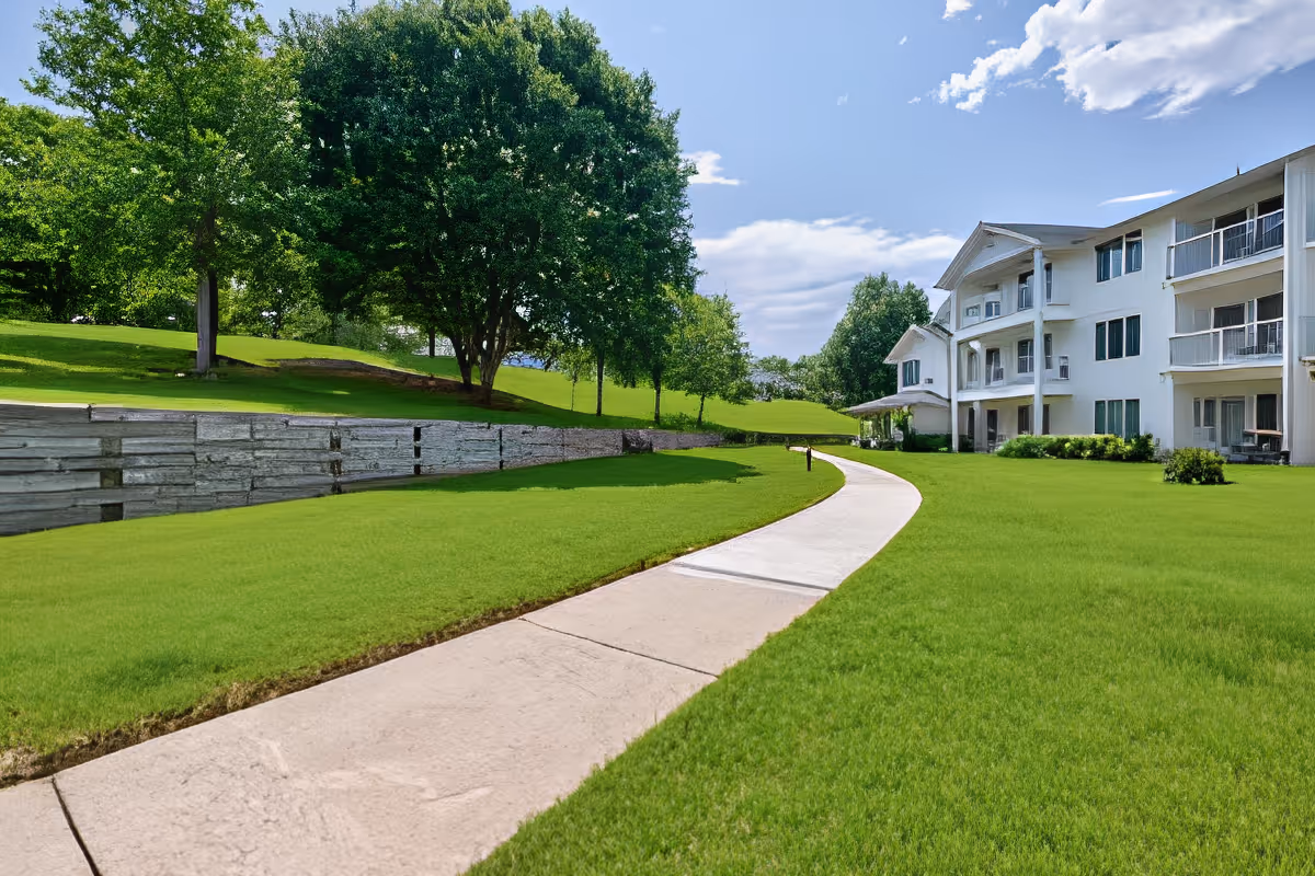 A curved concrete pathway runs through a well-maintained grassy area with trees and a retaining wall on the left. On the right side, there is a large white multi-story building with balconies and windows under a partly cloudy blue sky.