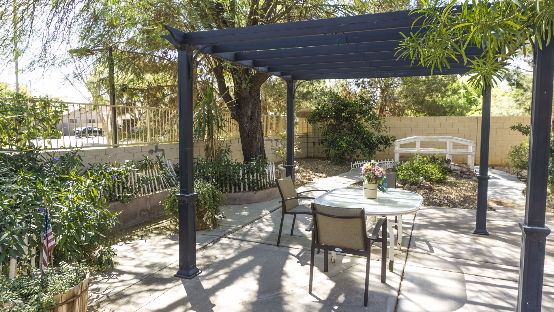 Outdoor patio area with a black pergola providing shade over a glass-top table and four chairs. The patio is surrounded by greenery including trees, bushes, and potted plants. A small white bridge and a beige brick wall are visible in the background.