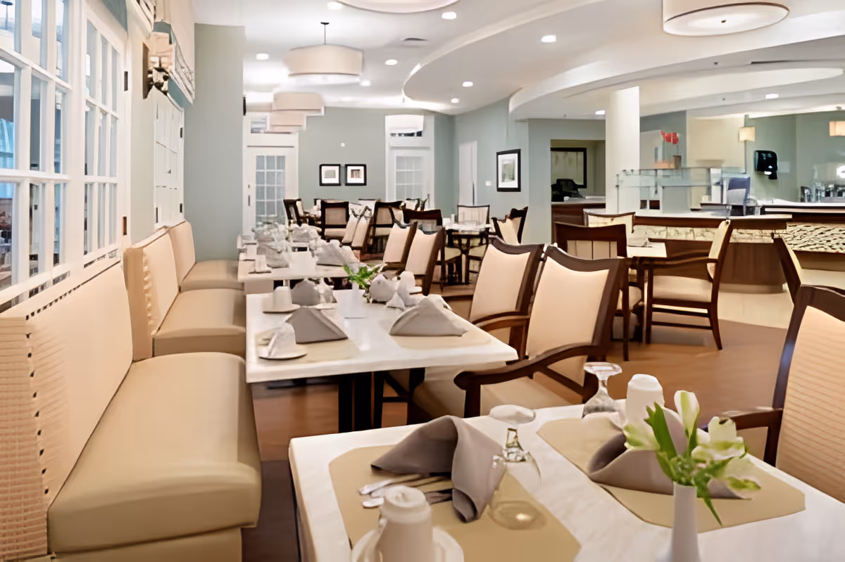 A bright and clean dining room with beige cushioned chairs and benches arranged around white tables set with napkins, glasses, and small flower vases. The room features soft lighting, light blue walls, and large windows on one side.