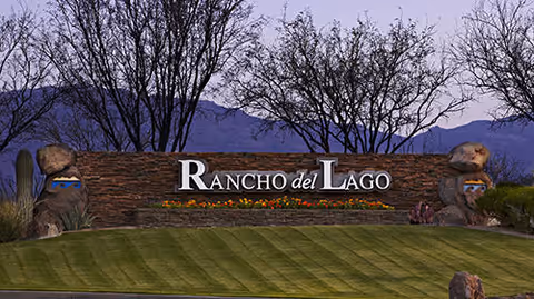 Entrance sign for Rancho del Lago with a stone wall background, decorative rocks on each side, and a well-maintained grassy area in front. Trees and mountains are visible in the background during dusk.