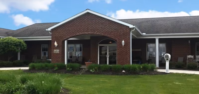 Front exterior view of a single-story brick building with a covered entrance supported by white columns, surrounded by green grass and shrubs under a blue sky with some clouds.