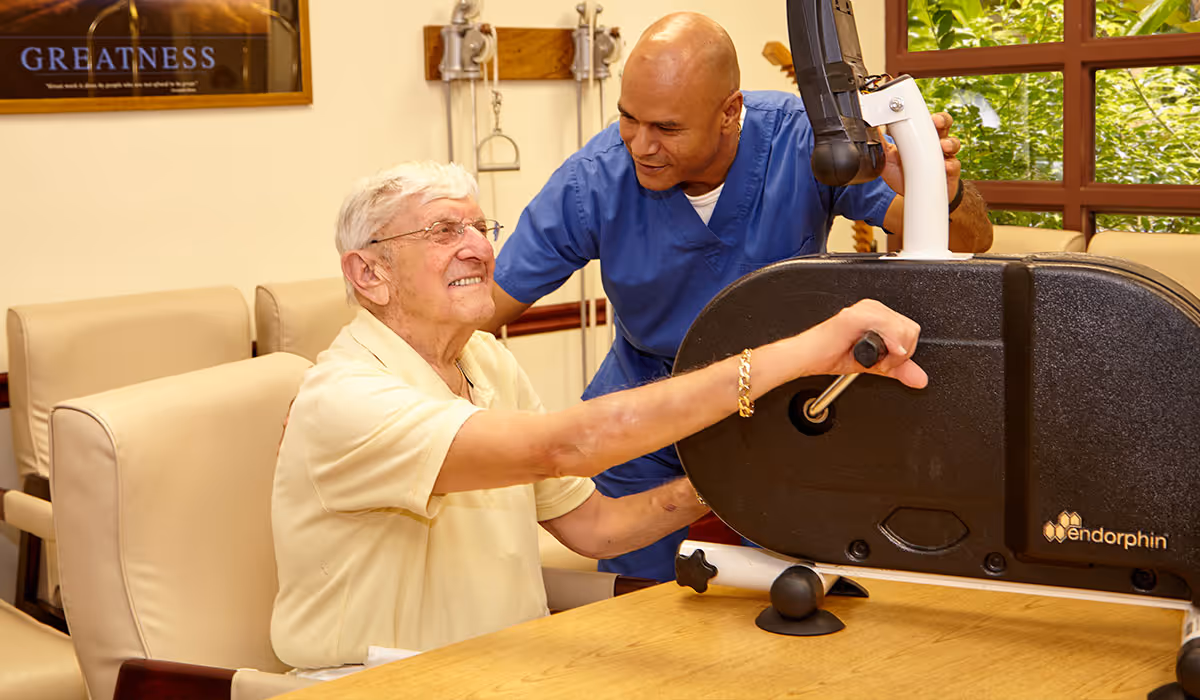 An elderly man in a yellow shirt uses a hand exercise machine while a caregiver in blue scrubs assists and encourages him in a well-lit room with beige chairs and a window showing greenery outside.