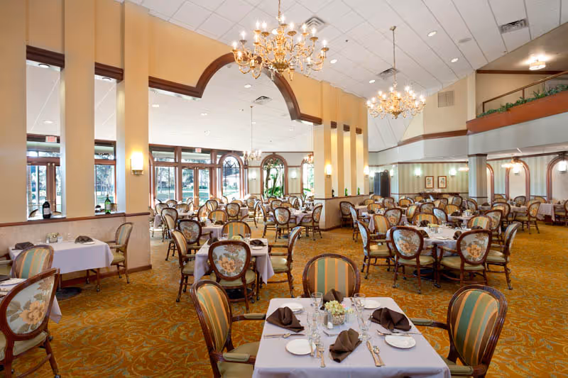Spacious dining room with multiple round tables covered in white tablecloths, set with plates, glasses, and brown folded napkins. The room features elegant chandeliers, large windows with views of greenery outside, and upholstered chairs with floral and striped patterns.