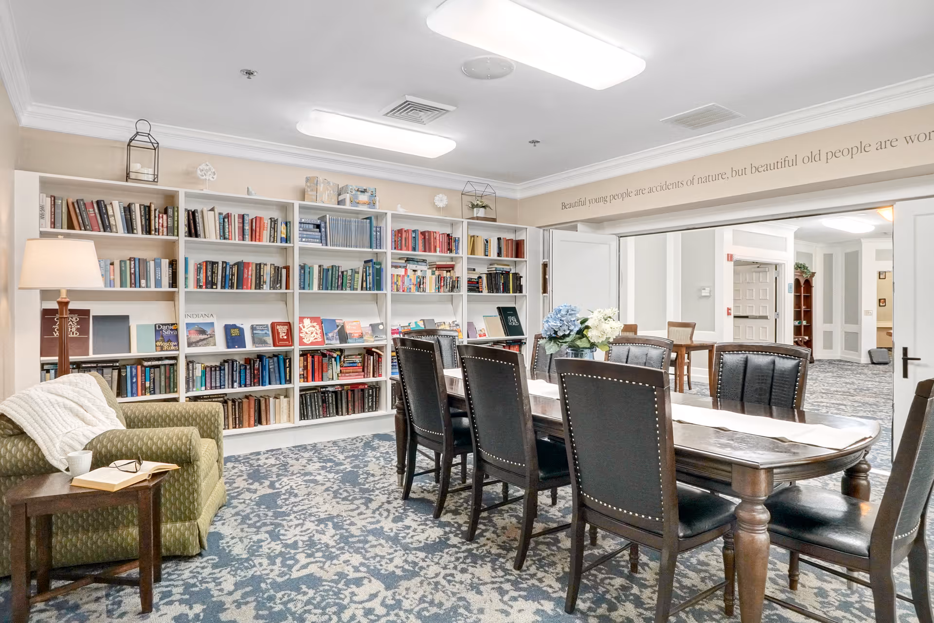 A cozy reading and meeting area in a senior living facility featuring a large wooden table with eight black leather chairs, a green armchair with a white throw blanket, a side table with a cup and an open book, and a wall of white bookshelves filled with books. The room has a patterned blue and white carpet and bright overhead lighting. Above the doorway, a quote reads, 'Beautiful young people are accidents of nature, but beautiful old people are wonderful works of art.'