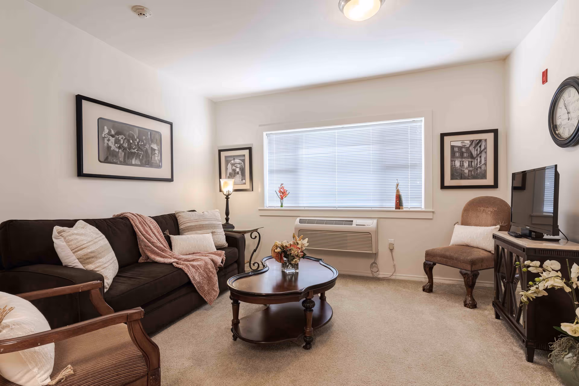 A cozy living room with a dark brown sofa adorned with beige and white pillows and a pink throw blanket. There is a wooden coffee table with a floral arrangement in the center of the room. A window with closed blinds is above a wall-mounted air conditioning unit. The room also features a wooden armchair with a white pillow, a brown upholstered chair, a small wooden cabinet with a flat-screen TV on top, and framed black and white artwork on the walls.