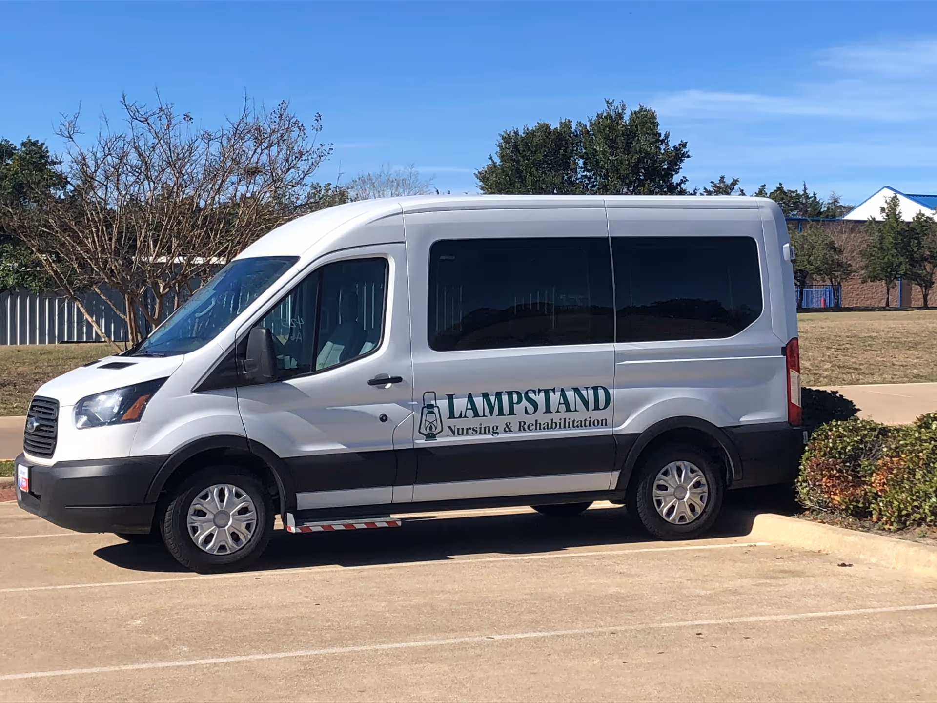 A white passenger van parked in a parking lot with the logo and text 'Lampstand Nursing & Rehabilitation' on its side. The background includes trees, a fence, and a building under a clear blue sky.