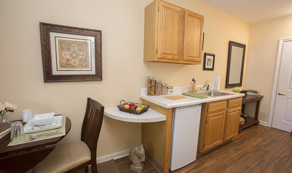A small kitchen area with wooden cabinets, a white countertop, a sink, and a mini refrigerator. On the countertop are containers with cereal, a soap dispenser, and a cutting board. To the left, there is a small dining table set with plates, glasses, and utensils, along with a chair. The walls are painted beige and decorated with framed artwork and a mirror. The floor is wooden.