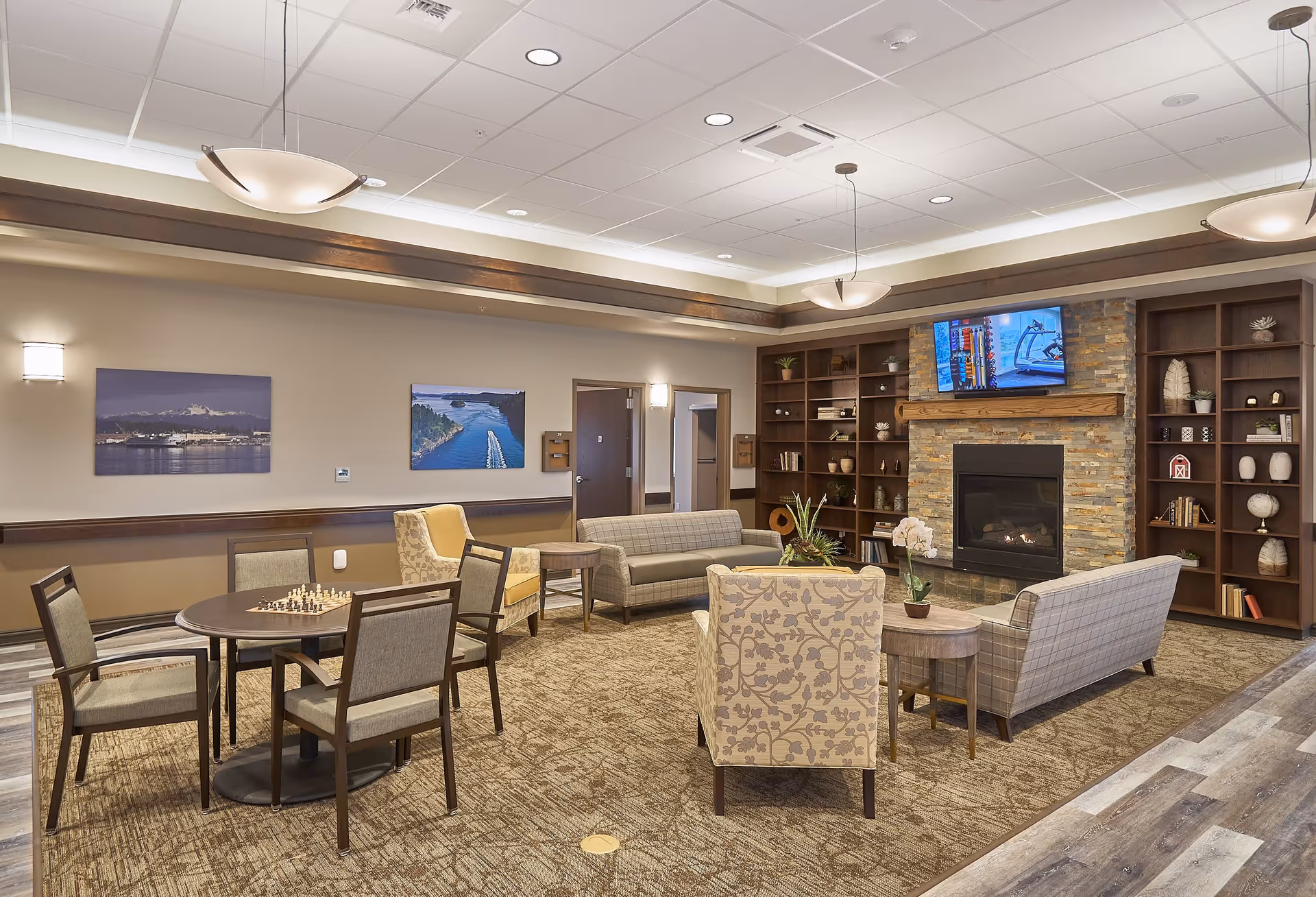 Comfortable senior living facility common room with seating around a fireplace, TV, bookshelves and a table with chairs.