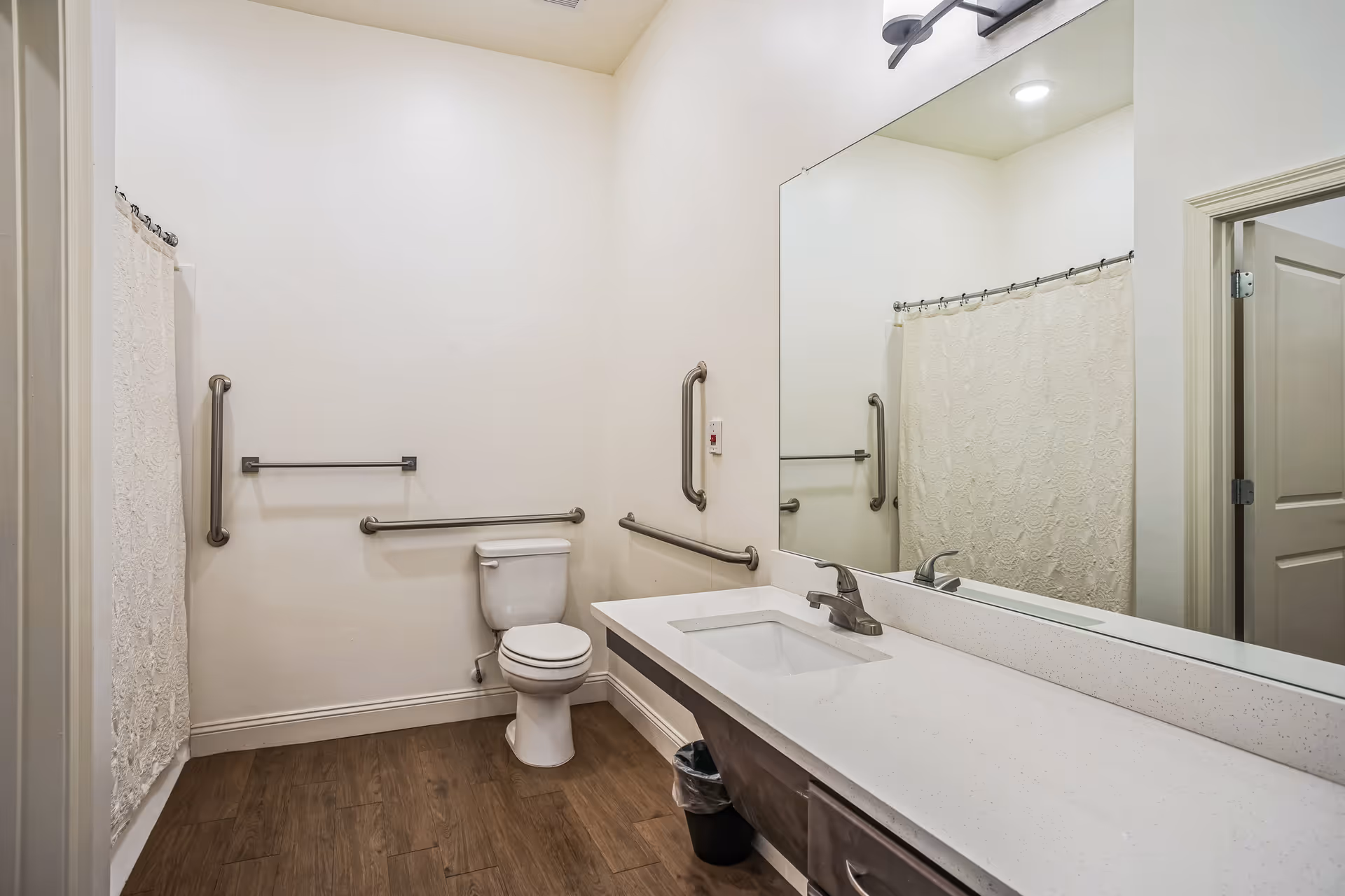 Accessible bathroom with a toilet, long vanity and sink, large mirror, grab bars, shower curtain, and wood-look flooring.