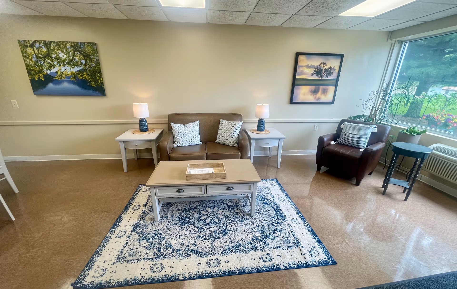 A cozy senior living room with a brown loveseat and two white patterned pillows, flanked by two white side tables each with a blue lamp. In front of the loveseat is a beige coffee table on a blue and white patterned rug. To the right is a brown armchair with a white pillow, a small black side table, and a large window showing greenery outside. The walls are beige with two framed landscape pictures hanging.