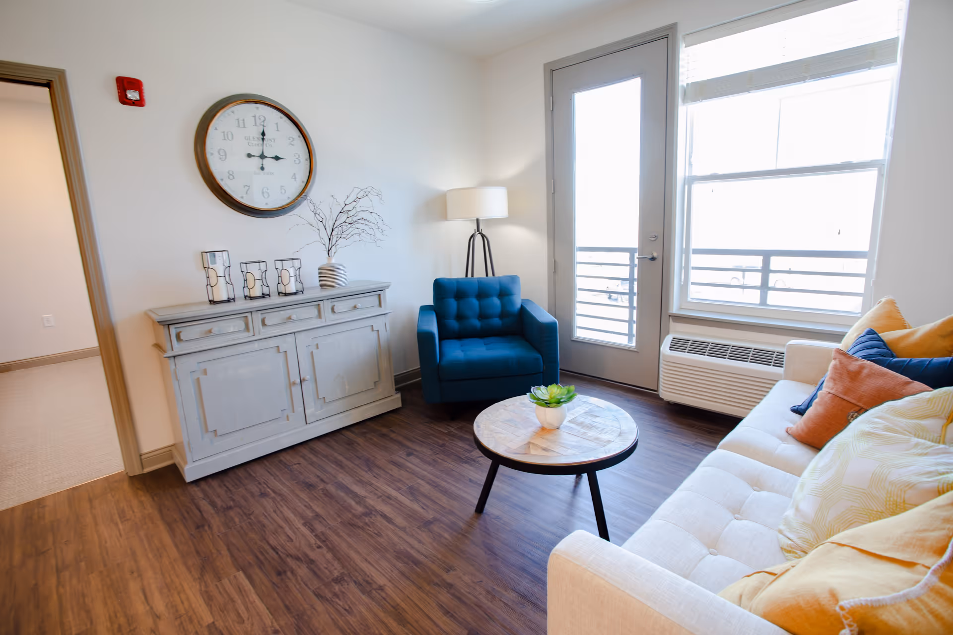 A bright living room with wooden flooring featuring a beige sofa with colorful cushions, a round wooden coffee table with a small plant, a blue armchair, a floor lamp, a white cabinet with decorative items, a large wall clock, and a glass door leading to a balcony with a window beside it.